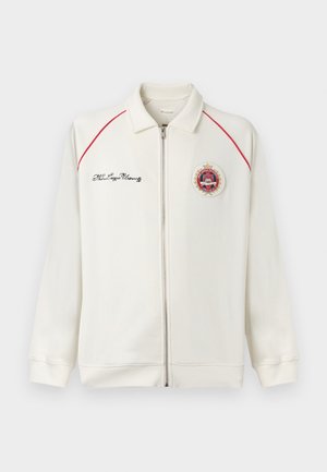 Veste blanche zippée avec passepoil rouge sur les épaules, inscription noire brodée sur la poitrine gauche et écusson rond sur la poitrine droite.