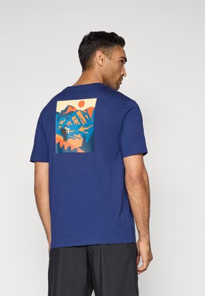 Mand iført blå t-shirt med farverig grafik af en person, der skitserer bjerge ved solnedgang på ryggen, parret med sorte shorts.