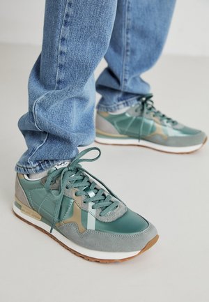 Person iført blå jeans og grøn-grå ruskind og læder sneakers med gummisåler, stående på et lyst gulv.