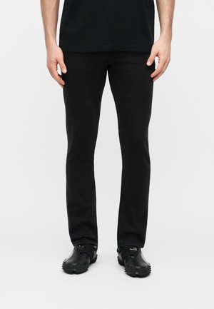 Homme portant un pantalon noir ajusté et des baskets noires, debout devant un fond blanc uni.
