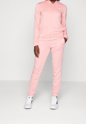 Femme portant un sweat à capuche zippé rose clair Adidas et un jogging assorti avec des baskets blanches Adidas, debout avec une main dans la poche.