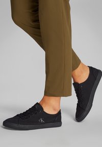 Isik, kes kannab lühikesi pruune pükse ja musti madalaid tossusid "cK" logoga, seisab hallil pinnal.