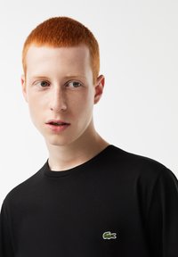 Camiseta de algodón negra con cuello redondo y mangas cortas. Presenta un pequeño logo bordado de un cocodrilo verde en el lado izquierdo del pecho.