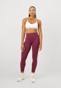 Sujetador deportivo blanco con una banda estampada, combinado con leggins burdeos de tiro alto. El modelo lleva zapatillas deportivas de color claro.