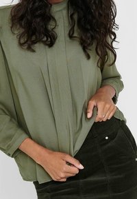 Vrouw met lang krullend haar die een losse olijfgroene blouse draagt, gestoken in een donkere corduroy rok, terwijl ze de zoom van de blouse met beide handen vasthoudt.