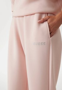 Šviesiai rožiniai sportiniai kelnės, pagamintos iš minkštos medžiagos, su šoniniu kišeniu ir sidabriniu uždaru "GUESS" logotipu priekyje. Lygus paviršius.