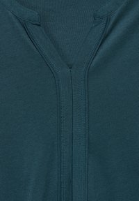 Türkises Baumwollmaterial mit V-Ausschnitt-Design, flachen Nähten und einer glatten Textur. Der Stoff wirkt dehnbar und angenehm zu tragen.