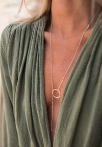 Gros plan sur une personne portant un haut texturé vert olive et un délicat collier lasso en argent avec un détail de boucle circulaire.