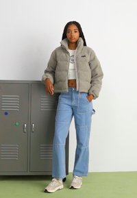 Giacca in pile grigia con zip e colletto alto, abbinata a pantaloni a gamba larga di un leggero blu e sneakers beige. Sullo sfondo semplice.