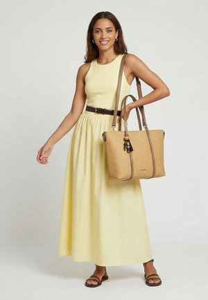 Donna sorridente in abito giallo senza maniche con cintura marrone e sandali, che tiene una borsa beige intrecciata con un portachiavi decorativo sulla spalla.
