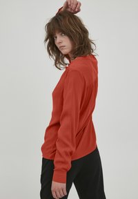 Blusa rossa a maniche lunghe con colletto abbottonato e dettaglio arricciato sul retro. Tessuto leggero con una superficie liscia, abbinata a pantaloni neri.