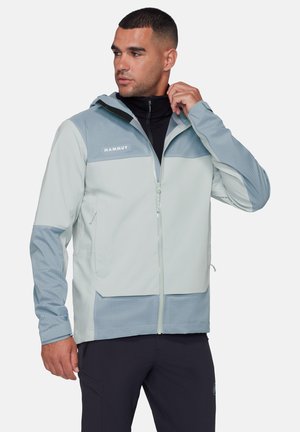 Homme ajustant le col d'une veste Mammut bleu clair et grise avec un pantalon noir, regardant vers sa gauche sur un fond uni.