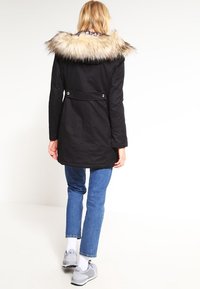 Svart parka med krage i fuskpäls, långa ärmar och midjebälte. Har två silverknappdetaljer på baksidan och är i knälång längd.