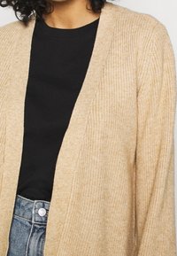 Beige stickad cardigan med ribbat mönster, öppen frontdesign och långa ärmar, lagd över en svart skjorta och högmidjade jeans.