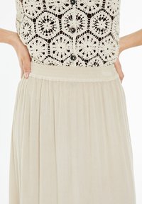 Falda plisada beige hecha de tela ligera con cintura elástica. Patrón texturizado visible, combinada con un top de ganchillo que presenta motivos hexagonales.