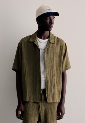 Ung mand iført en olivengrøn kortærmet skjorte med knapper og matchende bukser, hvid t-shirt med grafik, sølvkæder og tofarvet kasket.