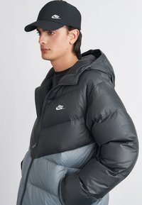 Sivi i plavi pufasti kaput s kapuljačom, s zatvaračem na zipper i Nike logom. Crna kapa s bijelim logom upotpunjuje odjevnu kombinaciju.