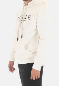 Sudadera crema con bolsillo frontal, puños elásticos y cordones. Presenta un diseño de texto negro en el pecho. Combinada con pantalones negros.