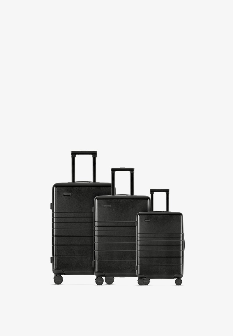 Drie zwarte koffers van verschillende maten, gemaakt van een hard materiaal, met horizontale groeven, uitschuifbare handvatten en wielen aan de onderkant.