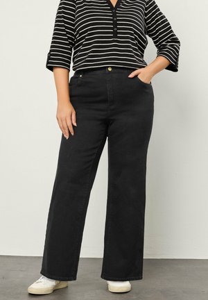 Person iført sorte bukser med vidde, en sort og hvid stribet langærmet skjorte og hvide sneakers, stående med den ene hånd i lommen.