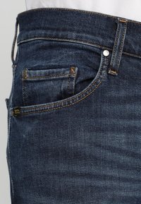 Närbild av mörkblå denimjeans med detaljerad orange sömnad, en framficka och en synlig liten myntficka.