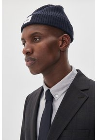 Tumes sinineustatud müts tekstuurse disainiga, millel on väike valge silt. Kandnud halli kraega särk ja tumedad mustrilised ülikonnad.