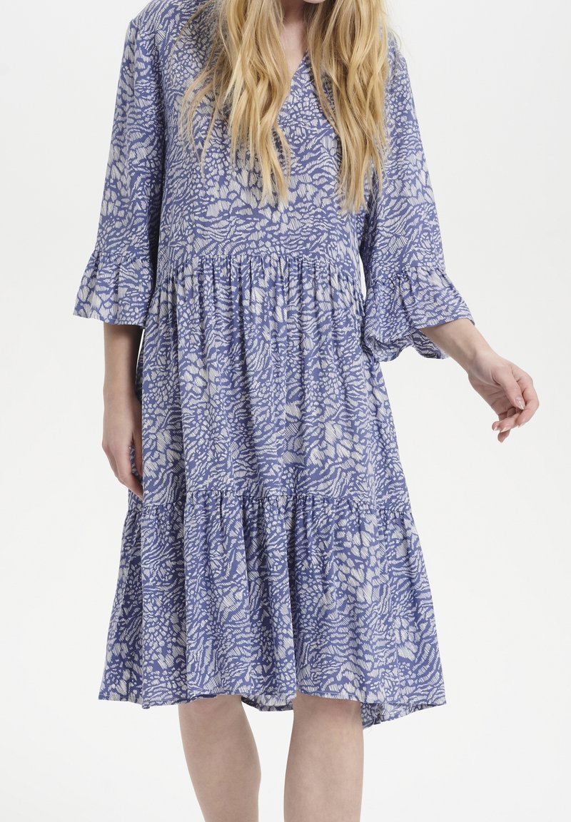 Abito blu a fantasia con scollatura a V, maniche a tre quarti a campana e orlo a strati con rouches. Il tessuto presenta un motivo floreale in tonalità più chiare.