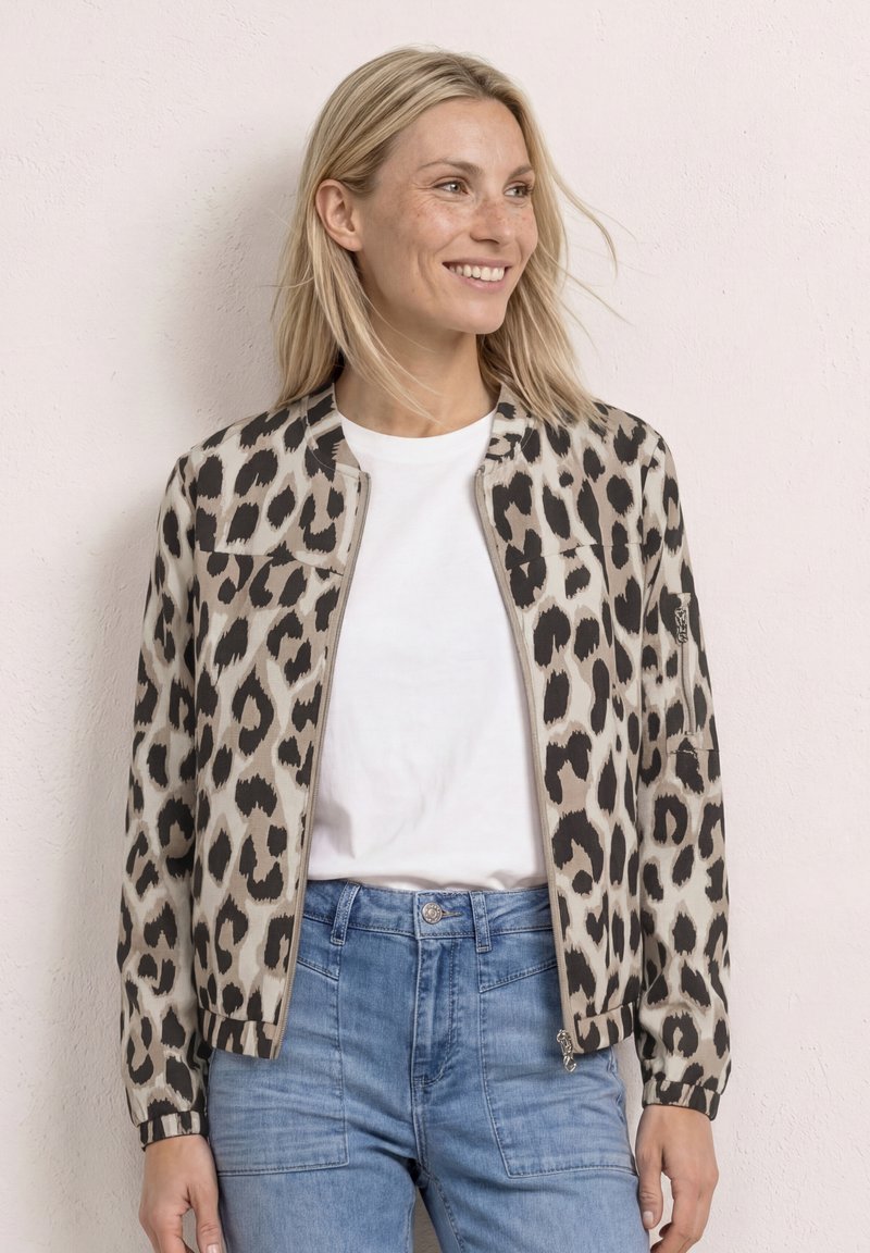 Smiling woman with blonde hair wearing a beige and black leopard print jacket over a white shirt and blue jeans, standing against a light wall.