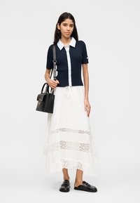 Žena v námorníckej modrej košeli na gombíky s bielym golierom, dlhej bielej sukni s čipkovanými panelmi, čiernych mokasínach a nesie čiernu kabelku.