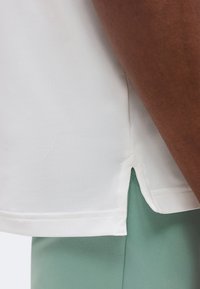 Gros plan de l'ourlet fendu sur le côté d'une chemise blanche portée sur un pantalon vert devant un fond uni.