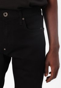 Jeans en denim noir au design ajusté, dotés d'un bouton en métal et de détails de poche. La texture paraît lisse avec un léger brillant.