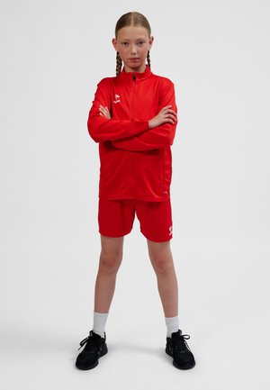 Red athletic tracksuit with zip-up jacket and shorts. Jacket features a fitted design, smooth texture, and logo. Worn with black sneakers.