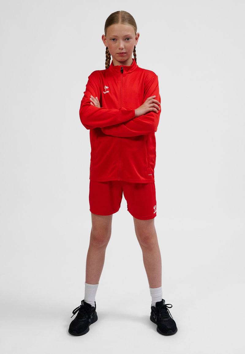 Tuta sportiva rossa con giacca con zip e pantaloncini. La giacca presenta un design aderente, una texture liscia e un logo. Indossata con scarpe da ginnastica nere.