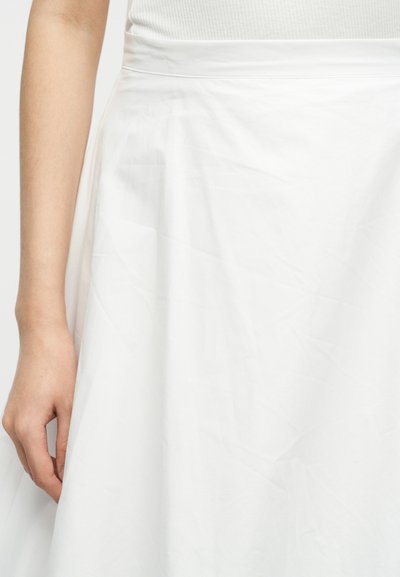 Γυναίκα που φοράει λευκή ψηλόμεση φούστα με λευκό ριπ τοπ, το αριστερό χέρι χαλαρό στο πλάι της, μπροστά σε απλό φόντο.