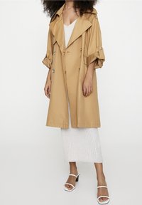 Lichtbruine trenchcoat met een dubbele rij knopen, oversized kraag en gathered mouwen, gecombineerd met een geribbeld witte jurk en witte sandalen.