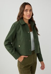 Donna con lunghi capelli castani che indossa una giacca verde corta con bottoni, una maglietta bianca e pantaloni verde oliva, con una mano in tasca su uno sfondo semplice.