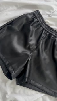 Melnas mākslīgās ādas šorti ar elastīgu jostasvietu, kas atrodas uz balta auduma virsmas.