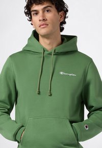 Giovane uomo con capelli ricci che indossa una felpa Champion verde con logo sul petto e sulla manica, mani nella tasca anteriore, sfondo neutro.
