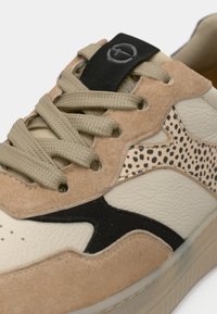 Gros plan d'une basket beige et crème avec des accents noirs, un détail imprimé léopard et des lacets beiges passés dans des œillets en daim.