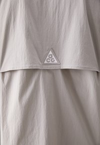 Light gray fabric top featuring a triangular ACG logo patch, smooth texture, and a loose design with a front flap detail.