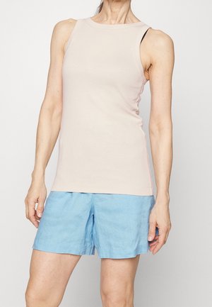 Kobieta w bezrękawnikowej beżowej bluzce i jasnoniebieskich szortach, stojąca na tle jednolitego białego tła.