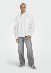 Witte blouse met lange mouwen en verticale blauwe strepen, voorzien van een borstzak, gecombineerd met lichtgrijze wijde jeans en grijze sneakers.