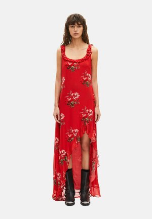 Kvinde iført en rød ærmeløs blomsterkjole med høj-lav skæringslinje og flæser, kombineret med sorte støvler til midt på læggen, stående mod en ensfarvet baggrund.