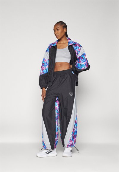 Čierny a viacfarebný teplákový komplet s kvetinovým vzorom, obsahujúci kratší kabát a zodpovedajúce nohavice. Viditeľné biele akcenty a logo.