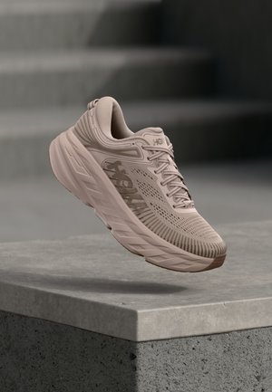 Scarpa da ginnastica beige con suola spessa e tomaia in mesh, sospesa sopra un bordo di cemento con gradini sfocati sullo sfondo.