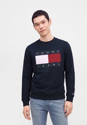 Νεαρός άνδρας που φοράει μαύρο φούτερ Tommy Jeans με κόκκινο και λευκό λογότυπο, σε συνδυασμό με ανοιχτό μπλε τζιν, στέκεται μπροστά σε απλό φόντο.