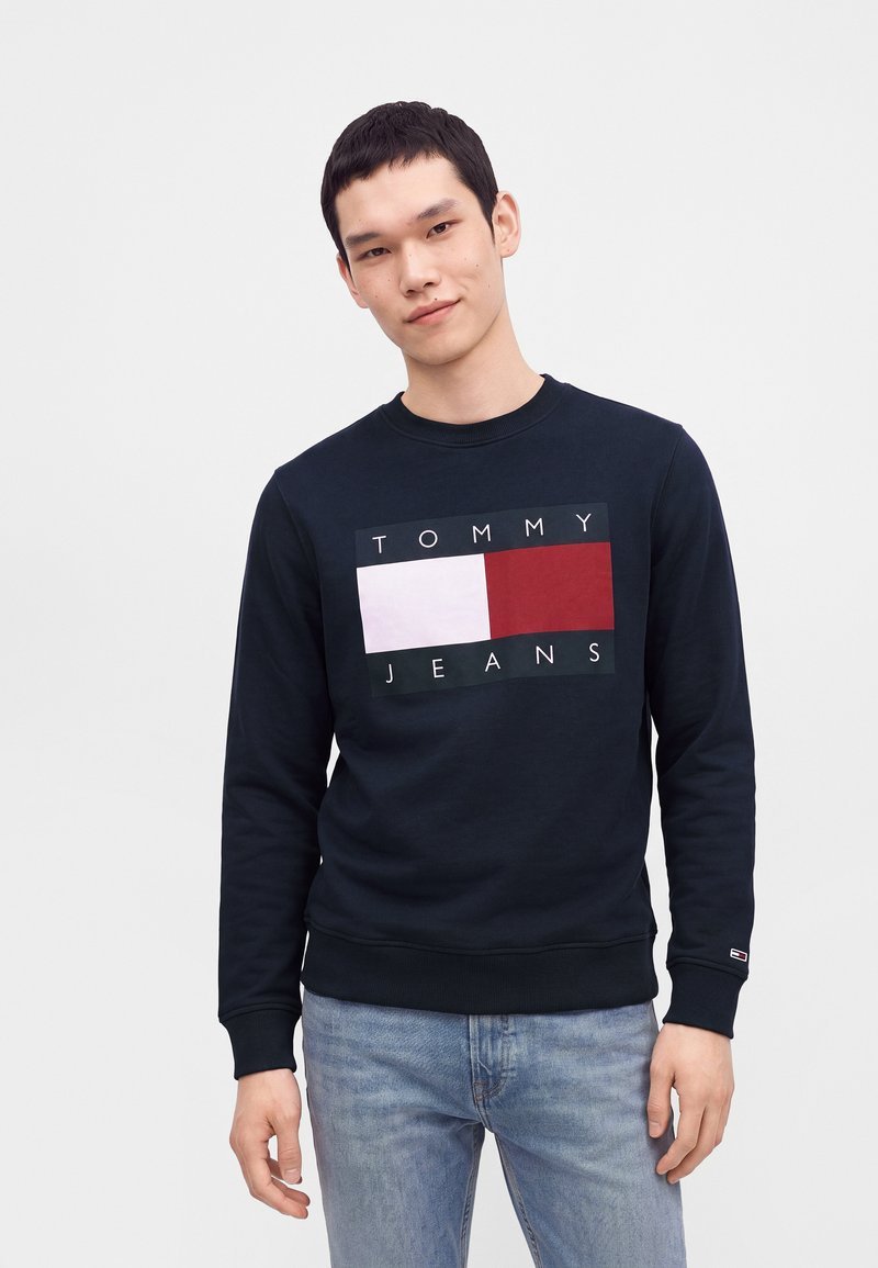 Νεαρός άνδρας που φοράει μαύρο φούτερ Tommy Jeans με κόκκινο και λευκό λογότυπο, σε συνδυασμό με ανοιχτό μπλε τζιν, στέκεται μπροστά σε απλό φόντο.