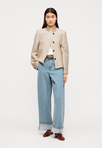 Γυναίκα που στέκεται φορώντας μια μπεζ ασύμμετρη σακάκι με κουμπιά, λευκό τοπ, φαρδιά τζιν παντελόνια με μανσέτες σε ανοιχτό γαλάζιο χρώμα, καφέ ζώνη και καφέ παπούτσια.