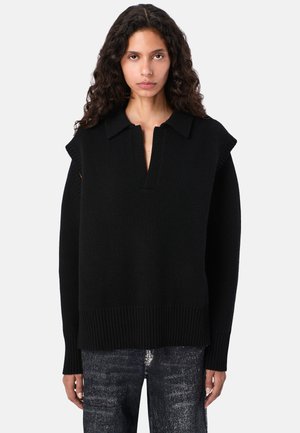 Frau mit lockigem Haar trägt einen schwarzen gestrickten ärmellosen Pullover über einem Langarmoberteil und dunkel gewaschenen Jeans, steht vor einem schlichten Hintergrund.