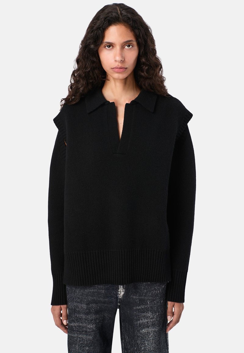 Kobieta z kręconymi włosami, ubrana w czarny dzianinowy bezrękawnik na długim rękawie oraz ciemne dżinsy, stoi na jednolitym tle.